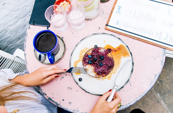 A woman at a restaurant eating blueberry pancakes with a cup of coffee and some water on her table